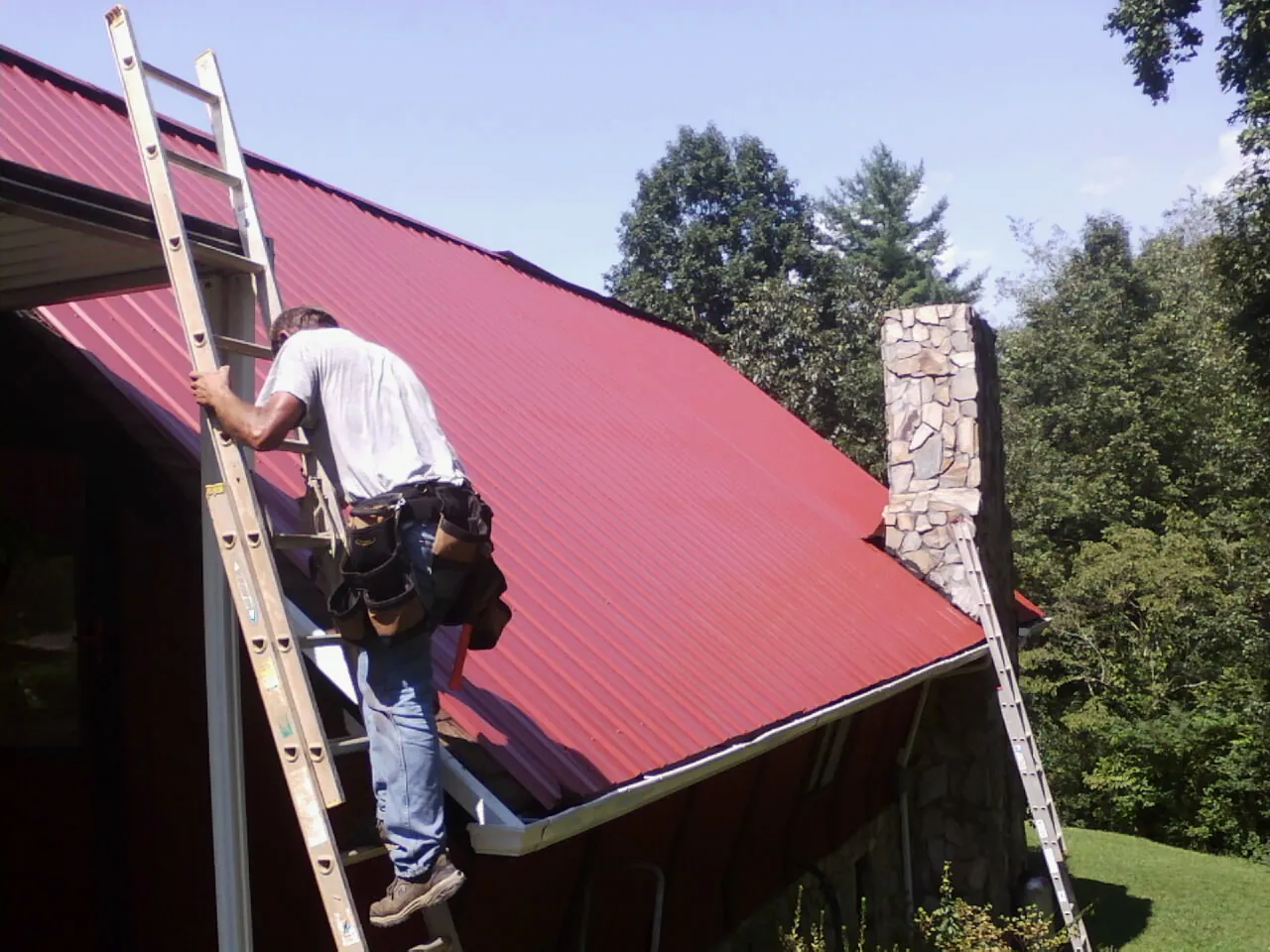 Roofer on a ladder beside a red metal roof and stone chimney — Soffit & Fascia Repair by Roofing Brothers in Horse Cave